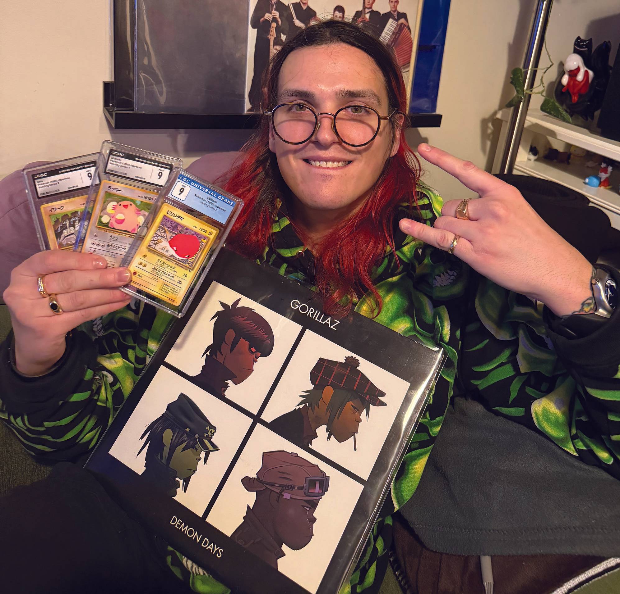 Person with red hair wearing a green hoodie holds Pok\u00e9mon cards and a Gorillaz album cover.