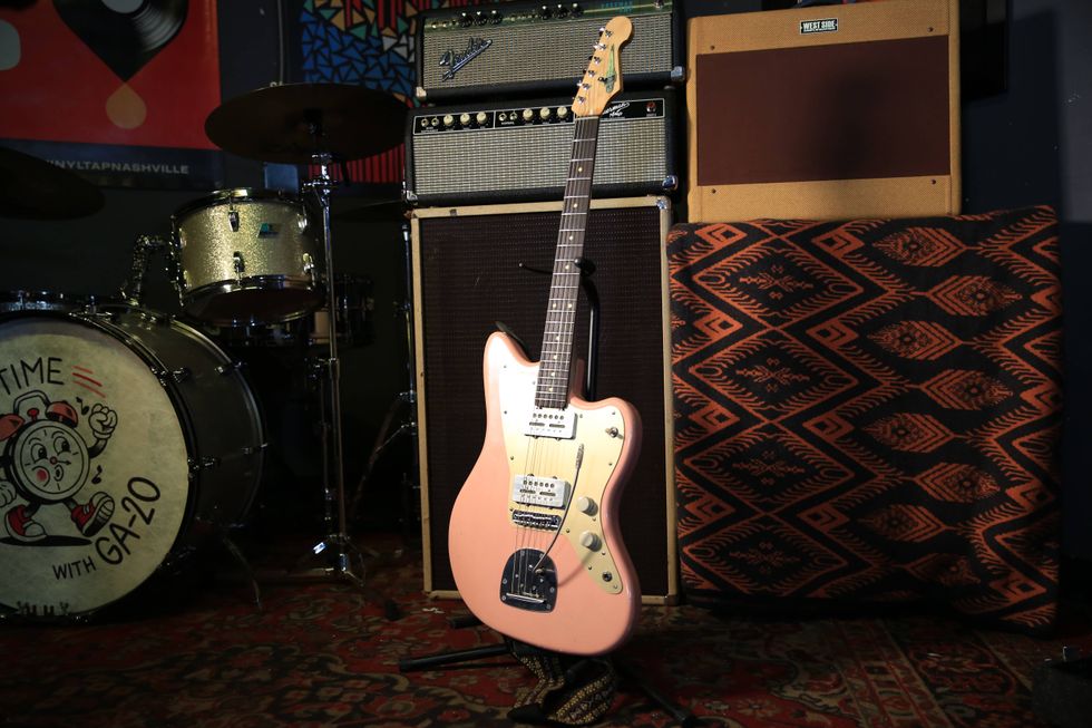 Pink electric guitar on stage beside a drum set and amplifiers in a music setting.