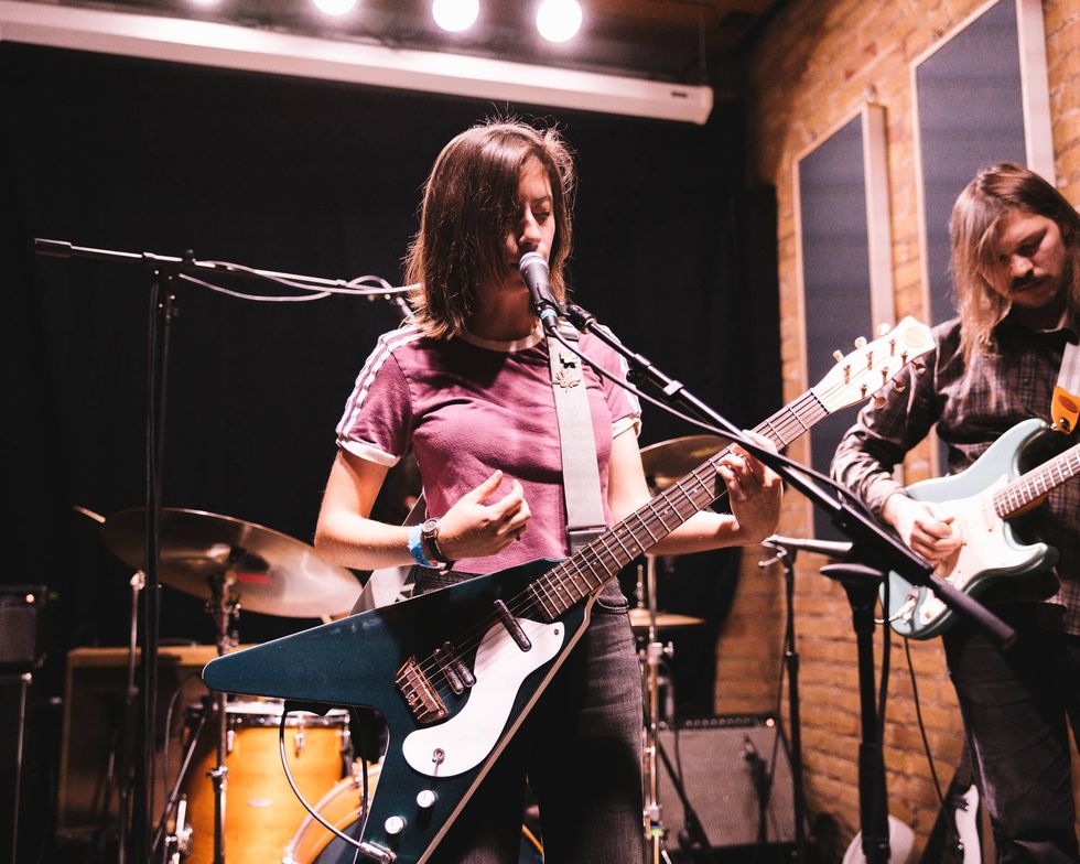 Singer performing with a blue guitar, backed by a drummer and another guitarist on stage.