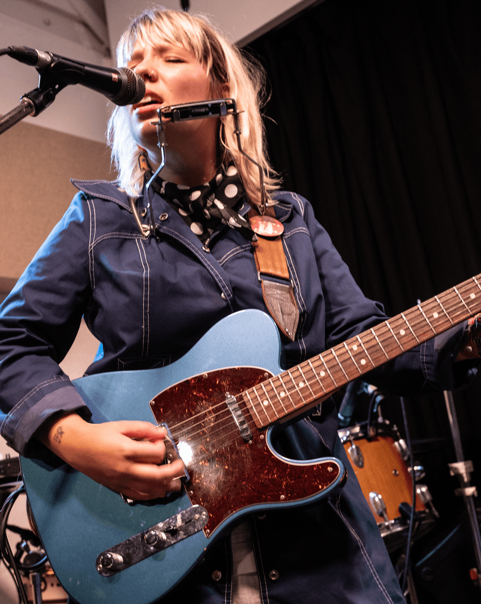 Singer-songwriter performs with a blue guitar and harmonica in a studio setting.