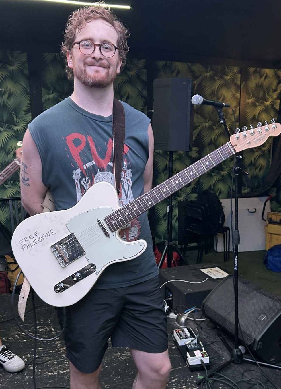 Smiling musician with curly hair holding a guitar, wearing a tank top with "Free Palestine" written on it.