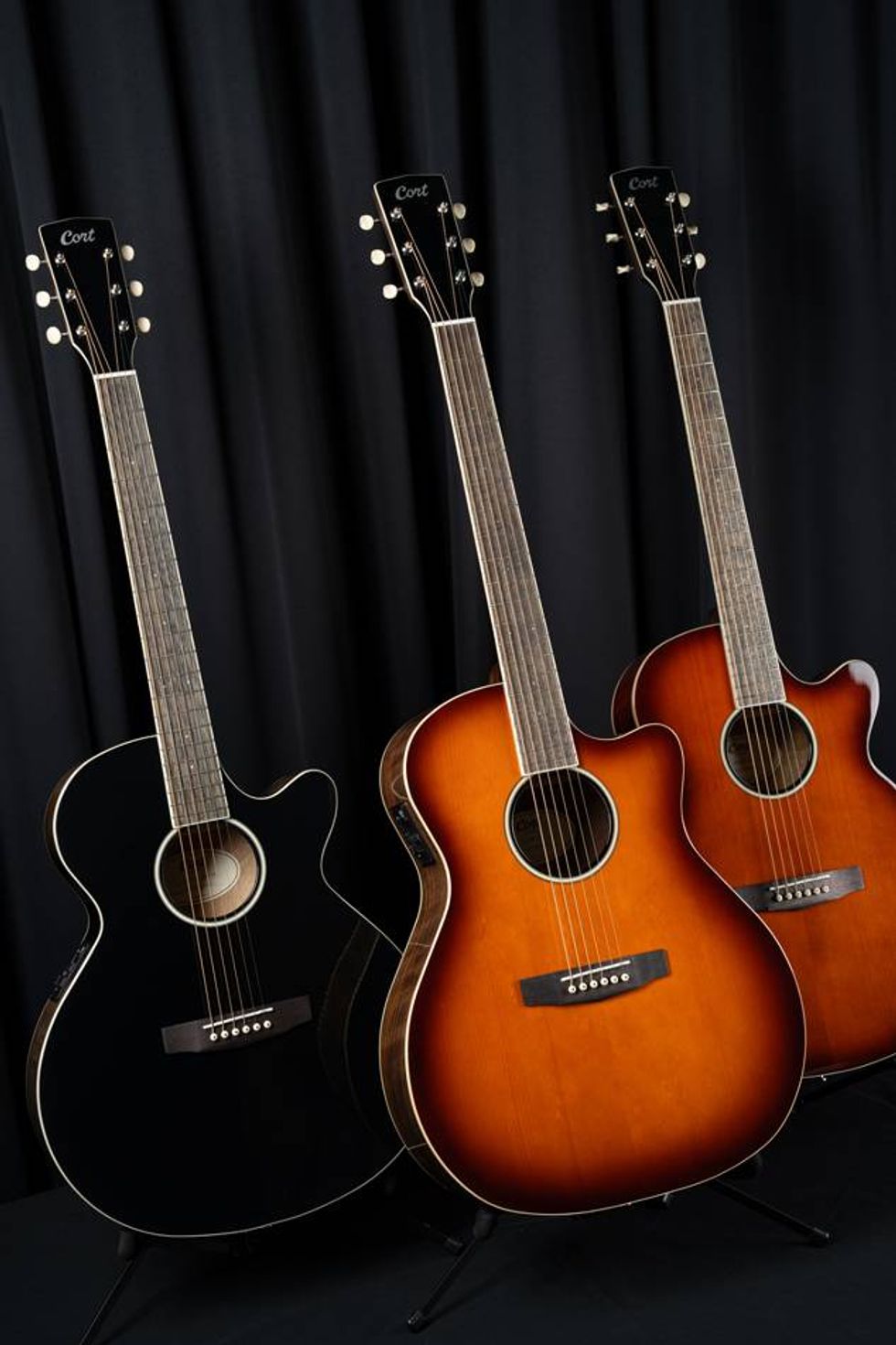Three Cort acoustic guitars in black, brown, and sunburst finish, displayed against a dark backdrop.