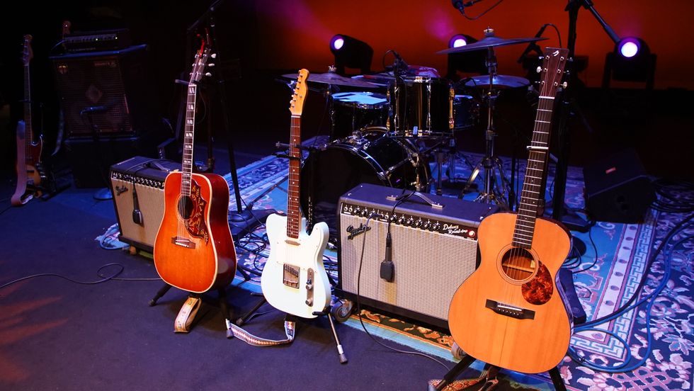 Three guitars on stage with amplifiers and a drum set in the background.