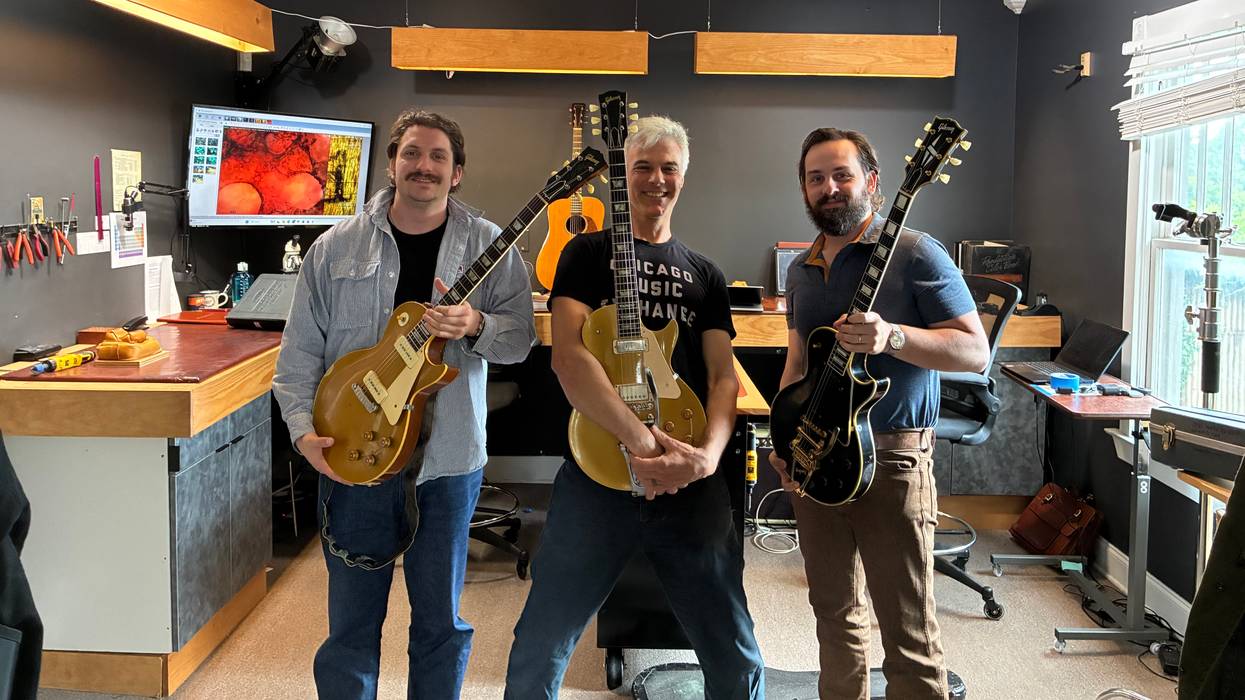 Three men hold electric guitars in a music studio, surrounded by tools and equipment.