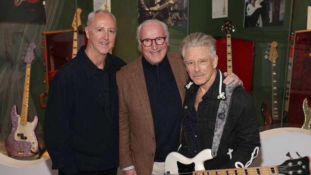Three men pose together in front of various guitars, smiling at the camera.