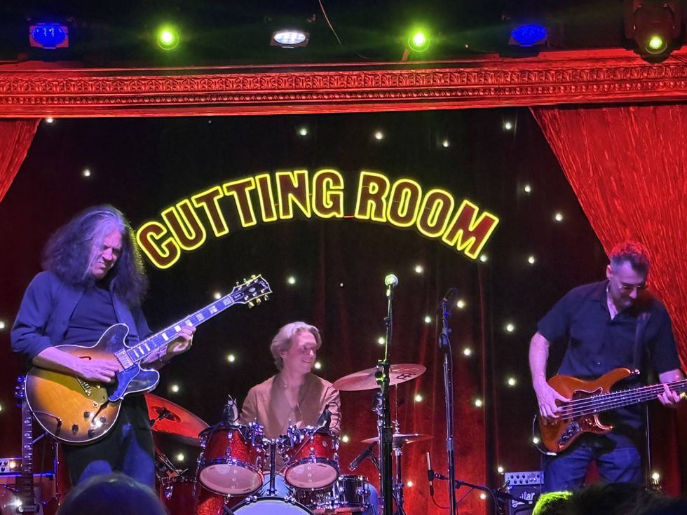 Three musicians perform on stage at the Cutting Room, illuminated by colorful lights.