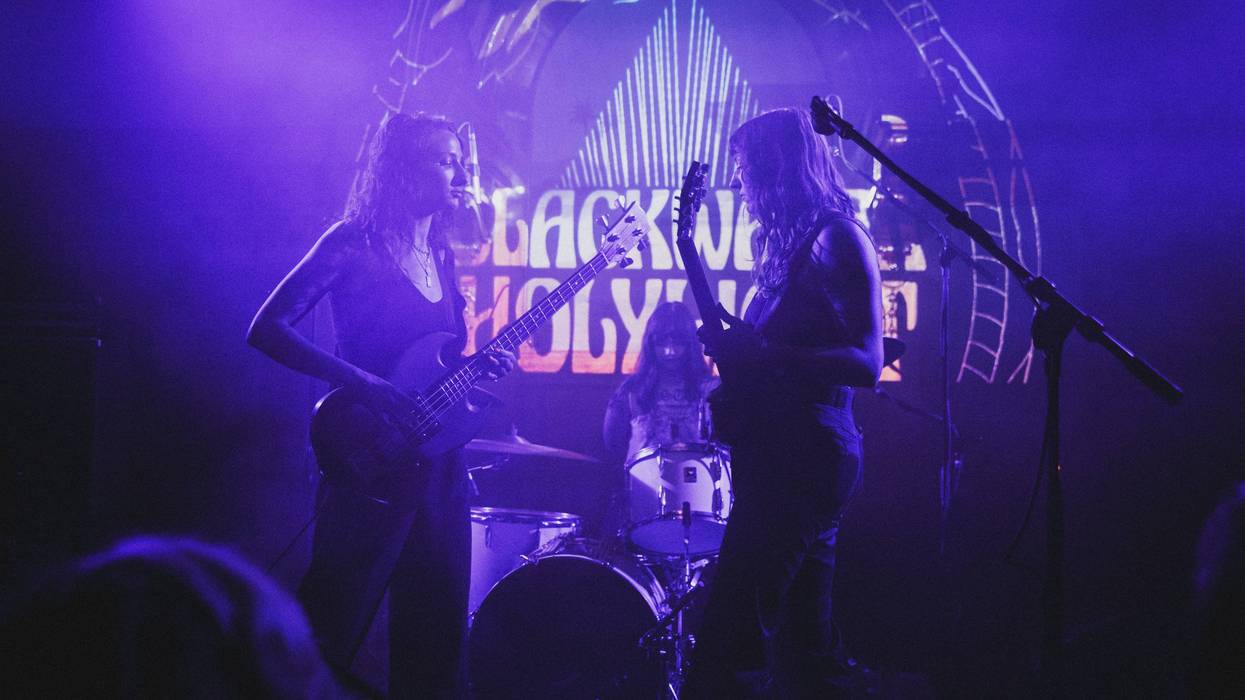 Three musicians perform on stage with vibrant purple lighting and a backdrop projection.