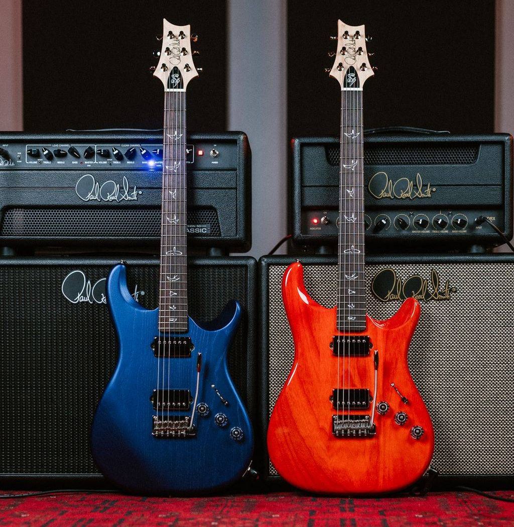 Two electric guitars\u2014a blue one and an orange one\u2014lean against amplifiers on a carpet.