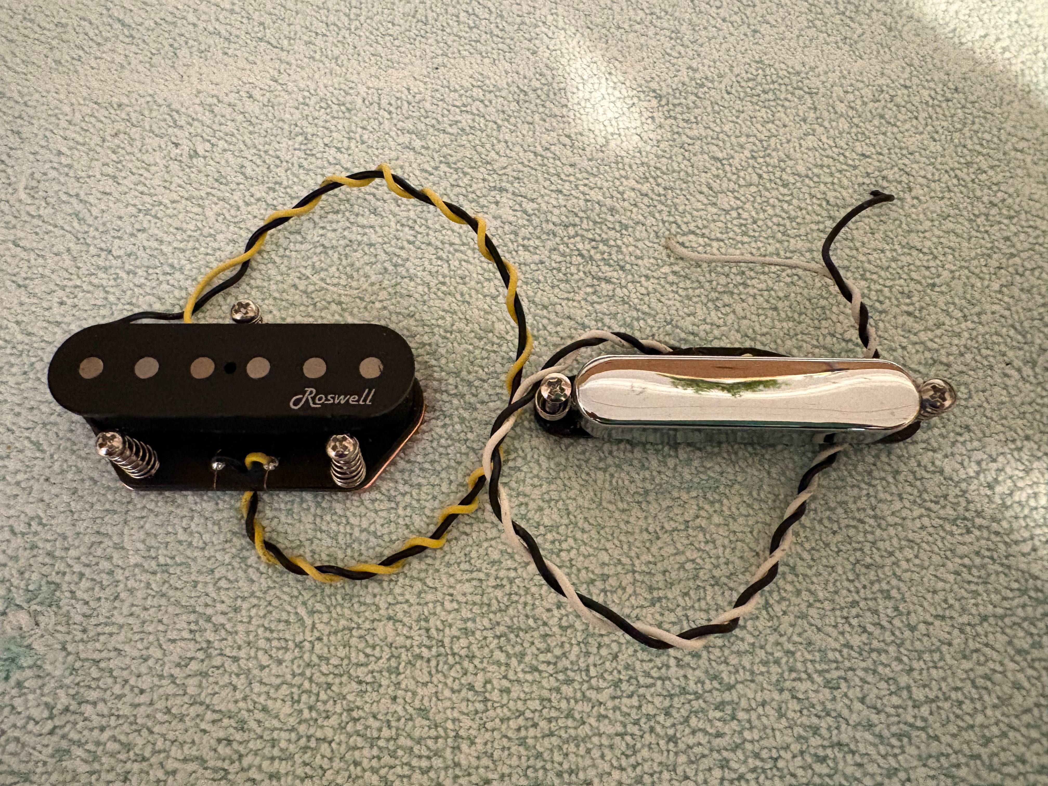 Two guitar pickups on a textured surface: one black with wires, one shiny silver with attached wires.