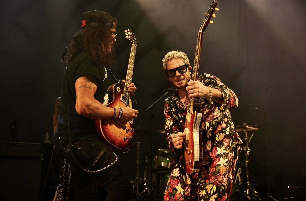Two guitarists perform energetically on stage, showcasing vibrant attire and instruments.