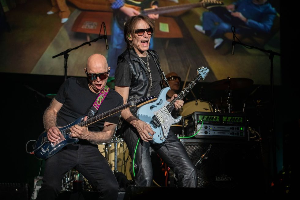 Two guitarists perform energetically on stage, with a lively audience and colorful lighting.