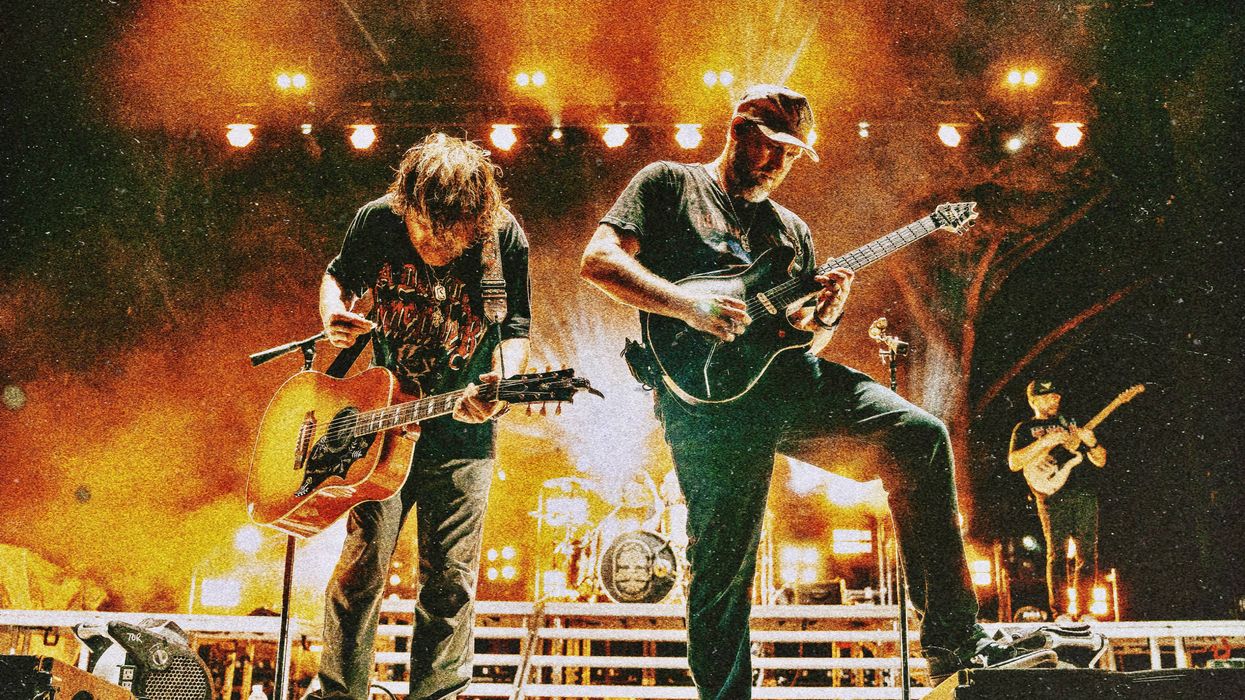 Two guitarists perform on stage, illuminated by vibrant lights, with a drummer in the background.