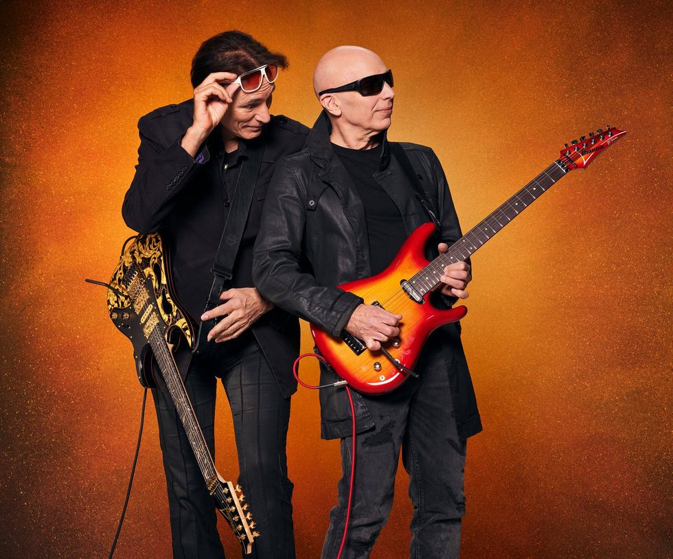 Two guitarists pose against a vibrant orange background, each holding an electric guitar.