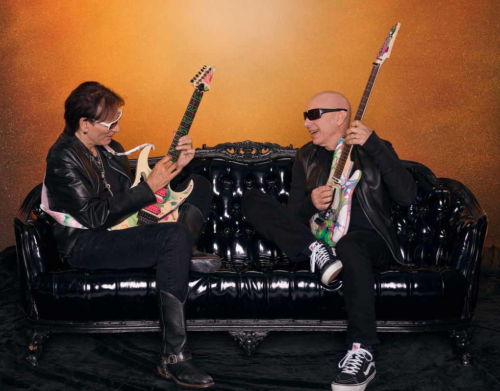 Two guitarists sit on a black couch, each holding colorful guitars, smiling at each other.