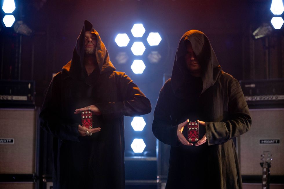 Two hooded figures hold devices against a backdrop of hexagonal lights and amplifiers.