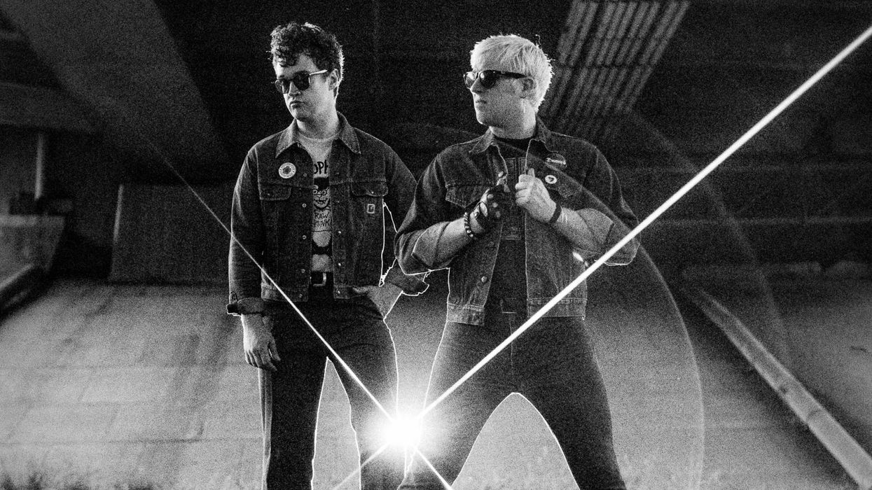 Two individuals in denim jackets stand confidently in a gritty, urban setting.