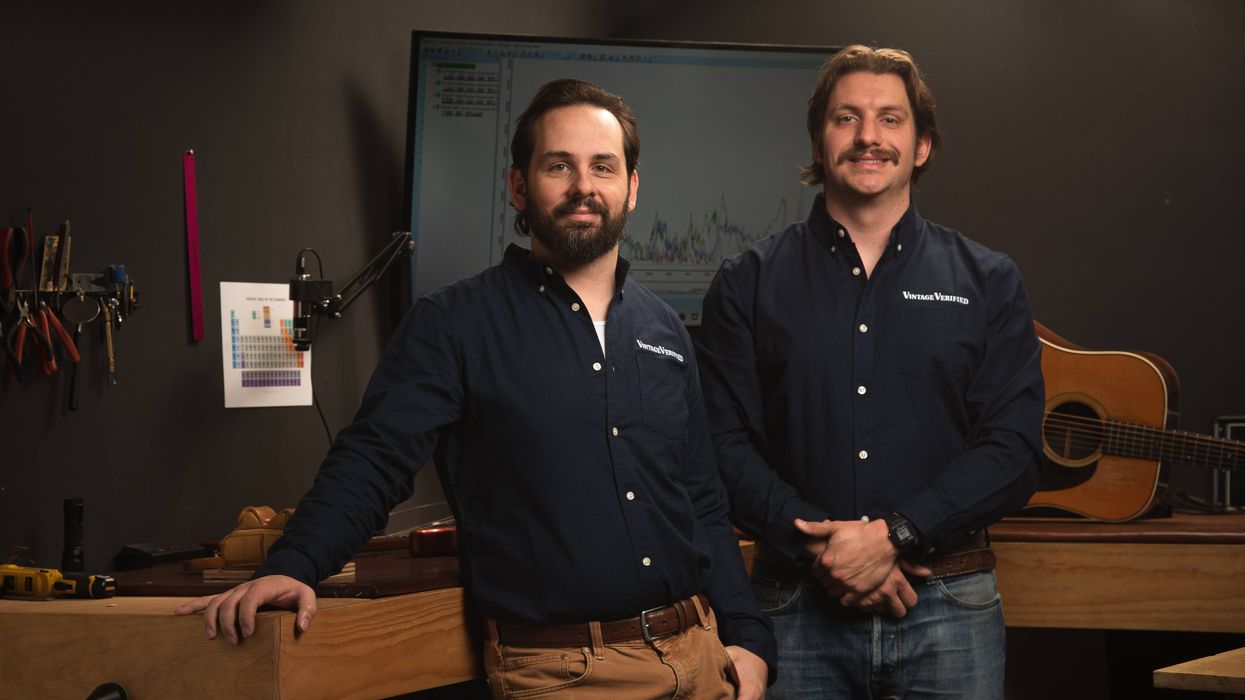 Two men in matching blue shirts stand in a workshop with tools and a guitar.
