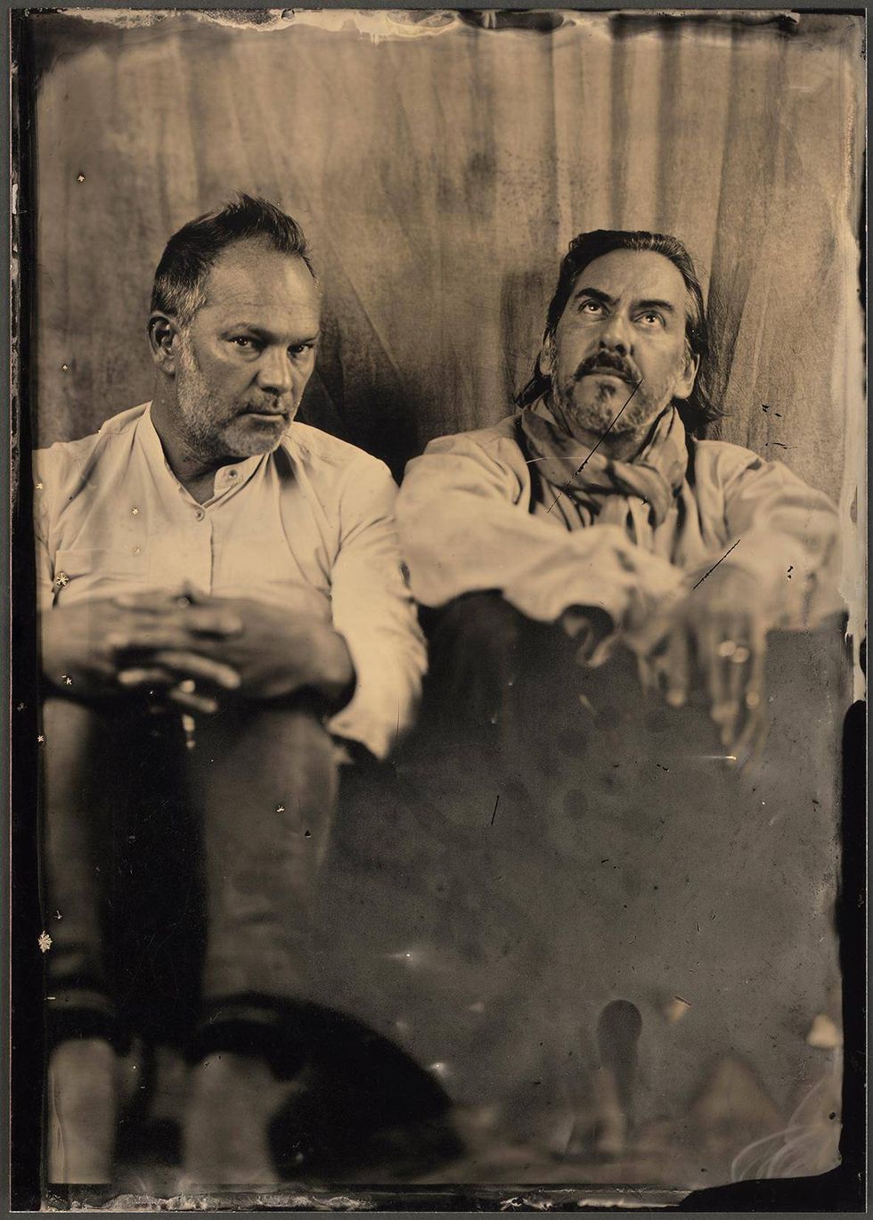 Two men in vintage clothing sit side by side, gazing thoughtfully in a dimly lit setting.