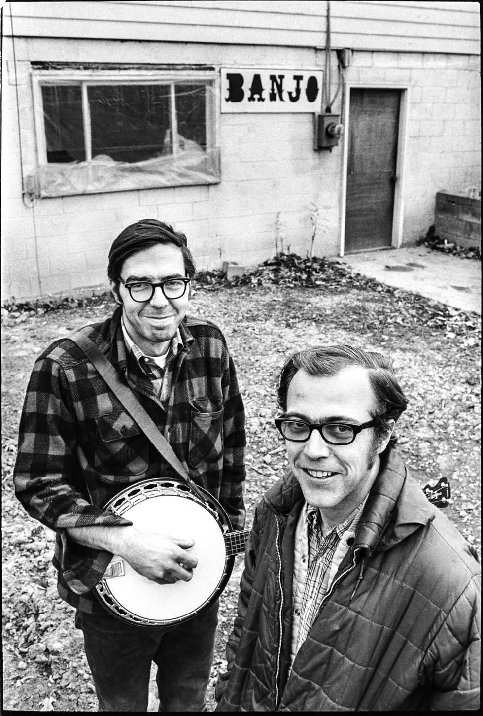 Two men, one holding a banjo, stand outdoors near a building with a "BANJO" sign.