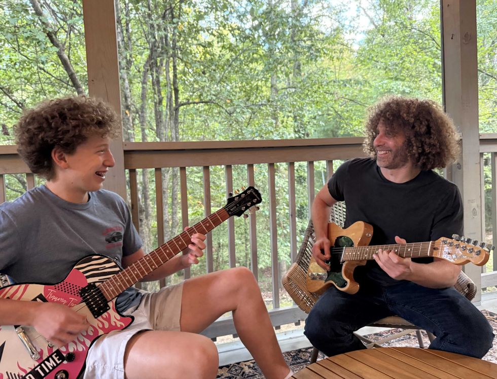 Two musicians joyfully playing guitars on a porch, surrounded by trees.