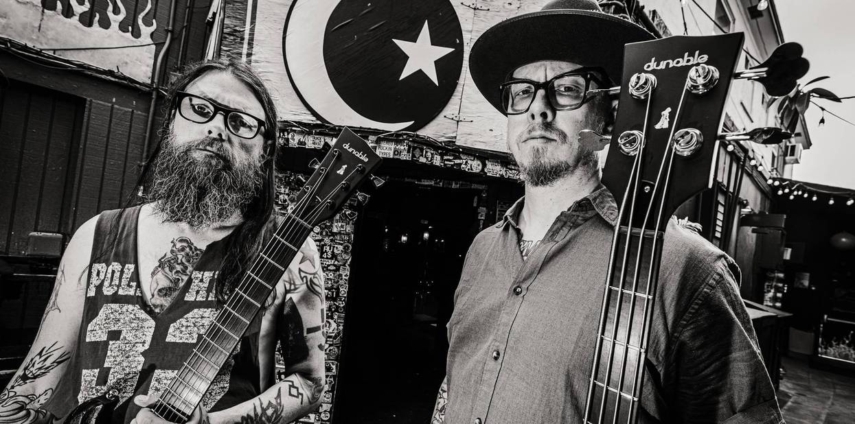 Two musicians, one with a guitar and the other with a bass, stand in front of a mural.