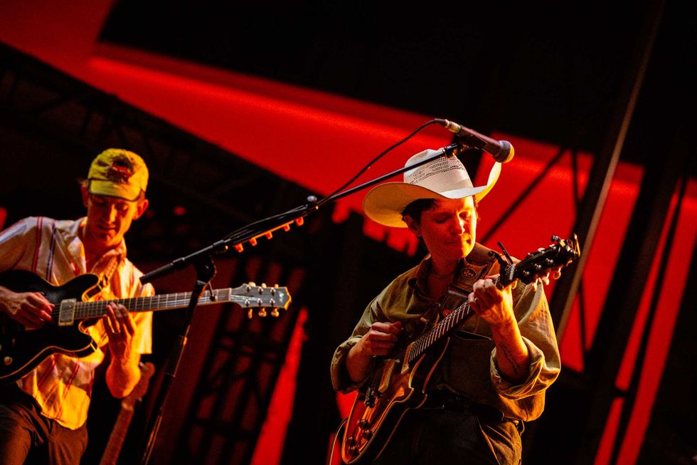 Two musicians perform on stage; one plays guitar while the other strums a mandolin.