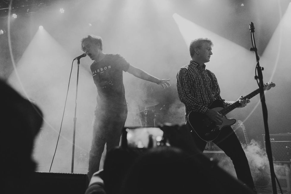 Two musicians perform on stage, surrounded by fog and dramatic lighting.