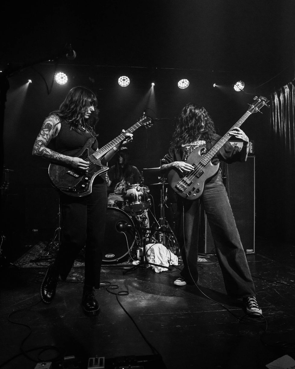 Two musicians performing on stage, one playing guitar and the other bass, with a drummer behind.