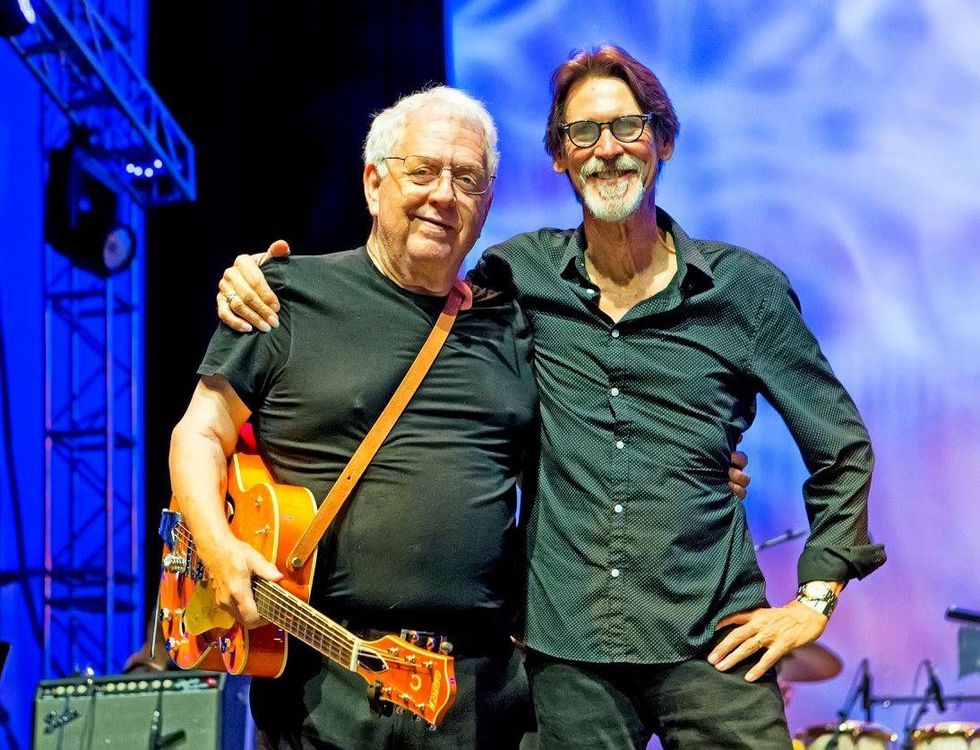 Two musicians pose together on stage, smiling; one holds an orange guitar.