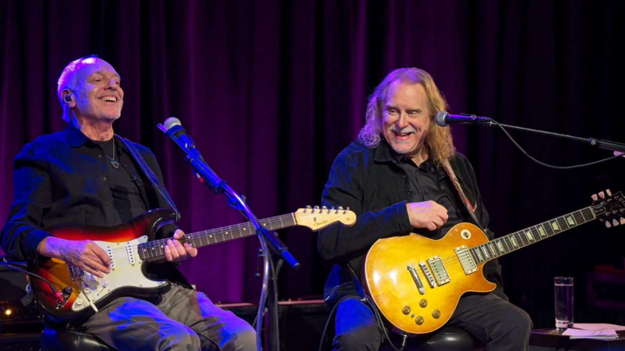 Two musicians smile and play electric guitars on stage, with a purple backdrop.