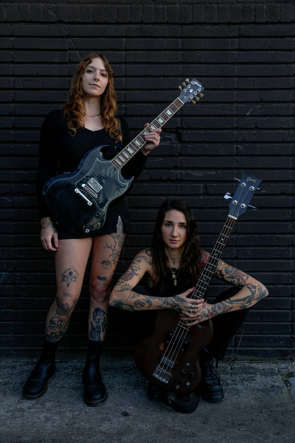 Two tattooed women pose with electric guitars against a textured black wall.