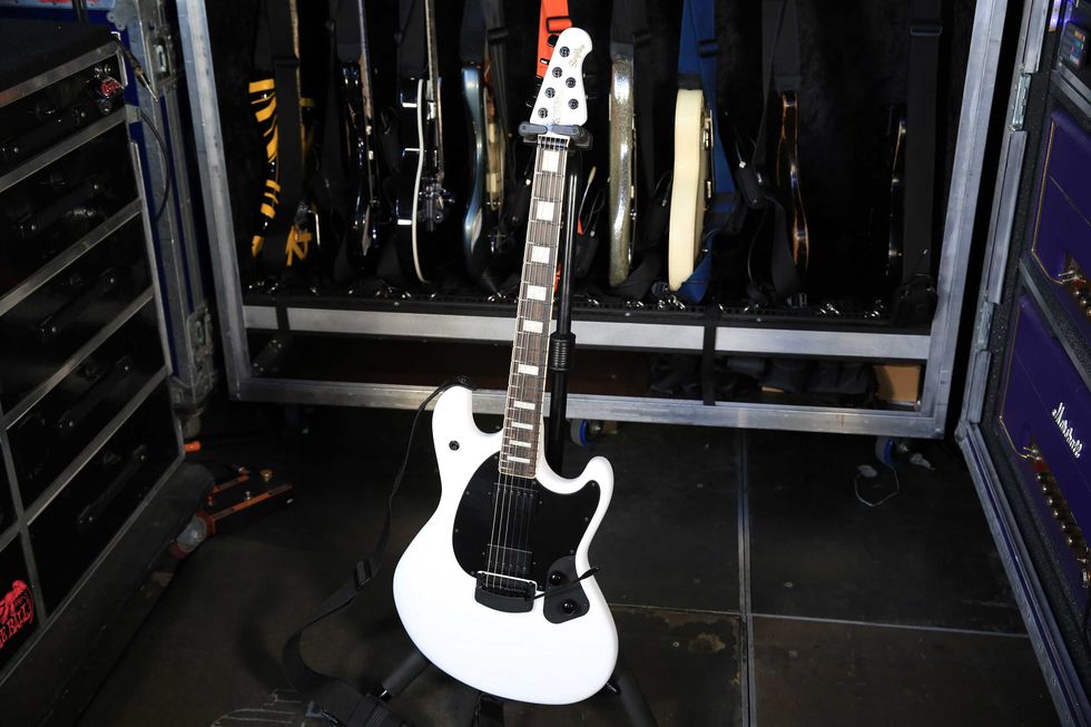 White electric guitar stands in front of a rack filled with various guitars.