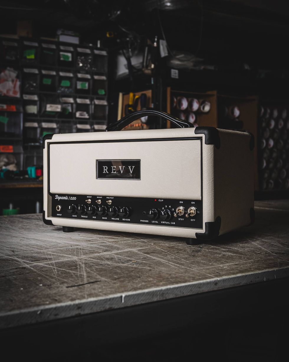 White guitar amplifier with knobs, labeled "REVV," on a wooden surface in a workshop.