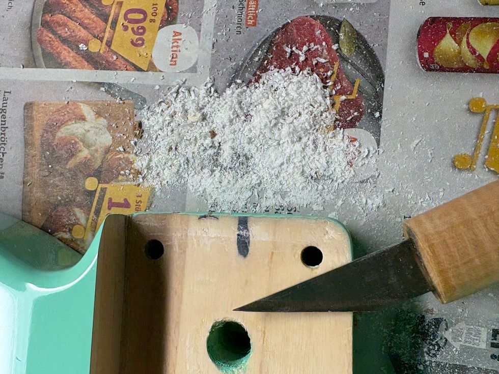 Wood shavings on newspaper next to a green wooden block and a knife cutting into it.