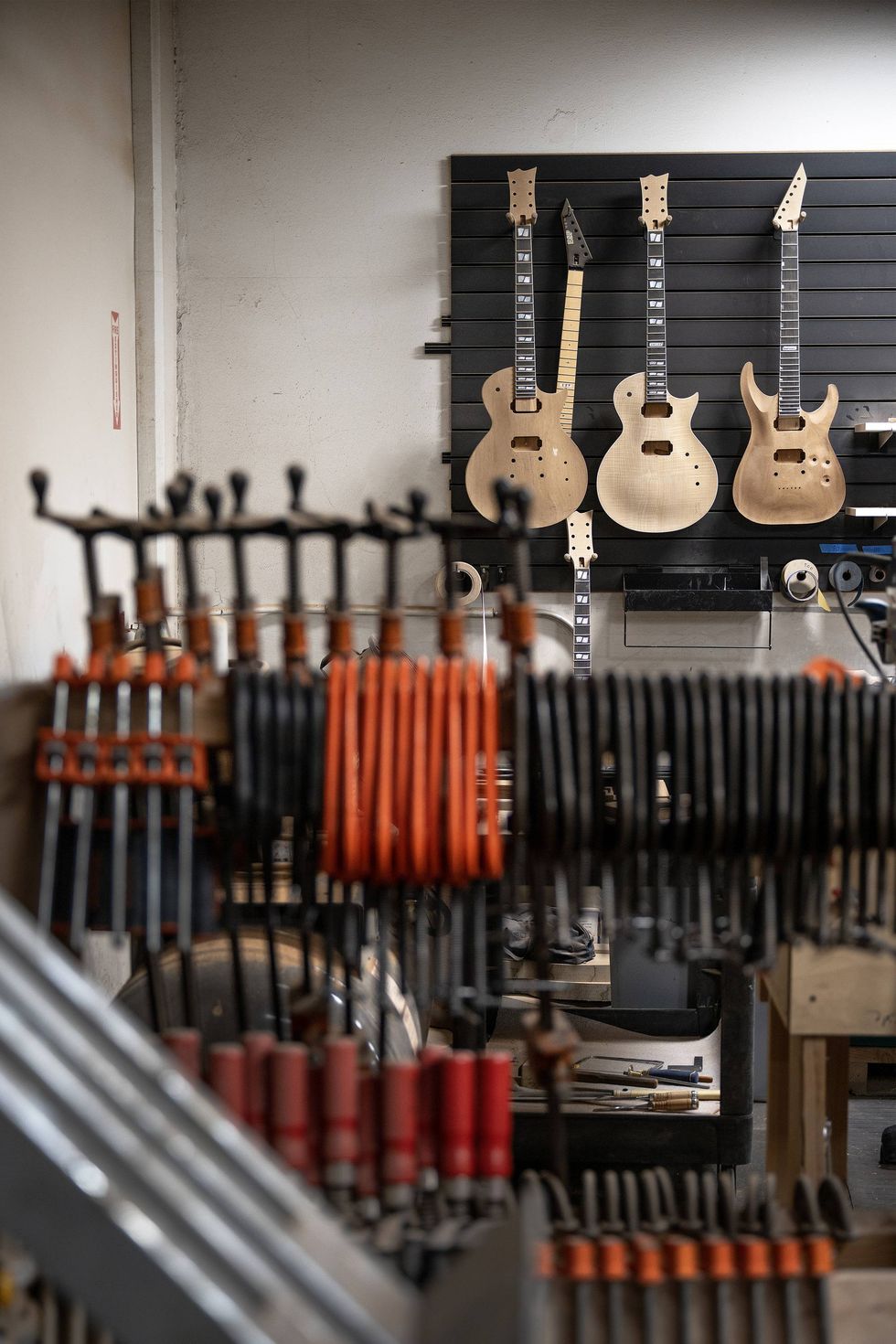 Wooden guitar bodies hang on a wall in a workshop, surrounded by various clamps.