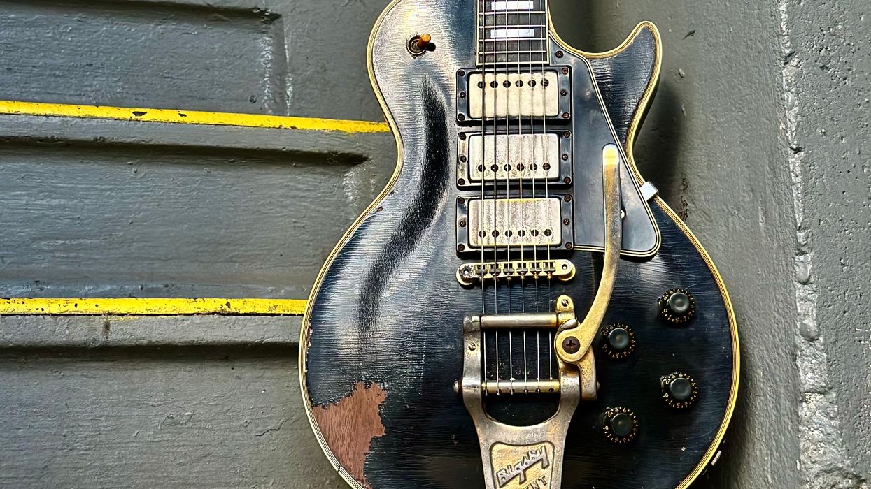 Worn black electric guitar hangs against a gray wall near yellow-striped stairs.